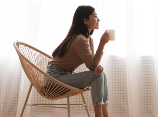 A person with a big smile is leaning forward in their chair as they hold a cup of coffee with both hands.