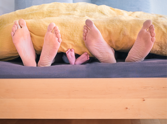 three pairs of feet are sticking outside of the blanket as a family lies in their bed together.