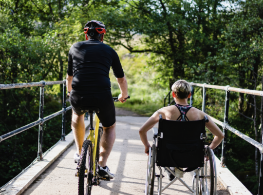 A person is steering their wheelchair forward on an outdoor trail and they are accompanied by an individual riding a bike.